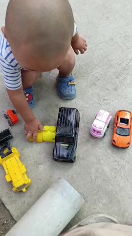 The video shows a young child in blue and white striped shirt and blue Crocs, crouching on a concrete surface. The child is playing with toy cars. In front of the child are several toy vehicles: a black SUV, a pink and white car, and an orange sports car. To the left of the black SUV is a yellow toy construction vehicle and yellow building blocks. Further to the left, on the ground, is a yellow toy car. In the background, a large blue toy car with pedals is visible. The child is reaching for a small orange toy car. The child moves the orange toy car. The child then picks up a small red toy car. The child is playing with the red toy car.
