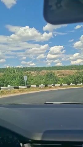 The video shows a view from inside a car looking forward and slightly to the right, as the car drives along a paved road. The road curves to the right, with guardrails on the left side. Beyond the guardrails, there is a landscape of green trees and rolling hills under a bright blue sky with scattered white clouds. In the distance, a person on a bicycle is visible on the side of the road. The car's dashboard is visible in the foreground.
