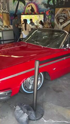 A red convertible is parked inside a building with graffiti art on the walls. In the background, several people are standing near a counter. The camera pans to the right, showing more of the graffiti art. Then, the scene shifts to an outdoor setting where a light green classic car is parked. A person's arm is leaning out of the driver's side window, and small flags are attached to the car.