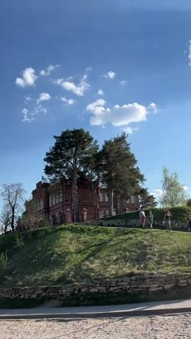 The video shows a view of a red brick building sitting atop a grassy hill. Several mature trees are situated around the building, partially obscuring it. A few people are walking along a path on the hill. The sky is blue with scattered white clouds. A low stone wall lines the base of the hill, and a sandy area is visible in the foreground.