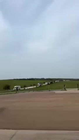 The video shows an open landscape with a paved foreground area that transitions into a green field. A road runs through the field in the distance, with several vehicles visible on it. The sky is overcast with a greyish-white hue. A wooden fence separates the paved area from the field. The vehicles appear to be a mix of cars and larger vehicles. There are no people visible in the frame.