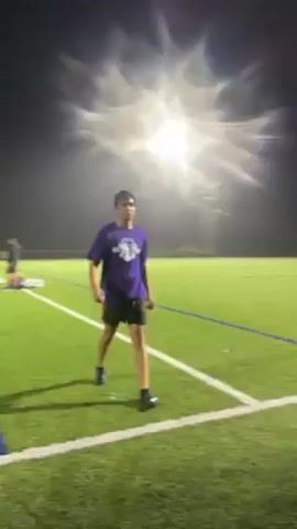 A young man wearing a purple t-shirt, black shorts, and black shoes walks over to a bench on a grassy field. He sits down and says in Spanish, “Que mierda de puto juego,” which translates to “What a piece of shit game.” He is illuminated by bright overhead lights. In the background is a soccer goal.