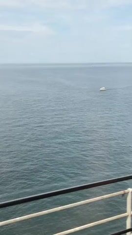A view from a vessel shows a calm, blue ocean under a cloudy sky. A small white boat is visible in the distance, moving from left to right. The foreground includes a railing, suggesting the perspective is from the deck of a larger ship.