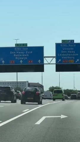 The video shows a section of a highway in the Netherlands, under a clear, bright sky. The camera is positioned from within a car driving on the highway, facing forward. Overhead road signs indicate directions: "RING-Oost" (Ring East) pointing straight ahead towards Den Haag-Zuid, Rotterdam, and Rijswijk via A4; and "RING-Noord" (Ring North) pointing left and right towards Utrecht, Den Haag, Leidschendam, and Voorburg via A12 and N14. Several cars and vans are traveling on the road in the same direction. Streetlights line the highway, and in the distance, there's a view of a building among some greenery.