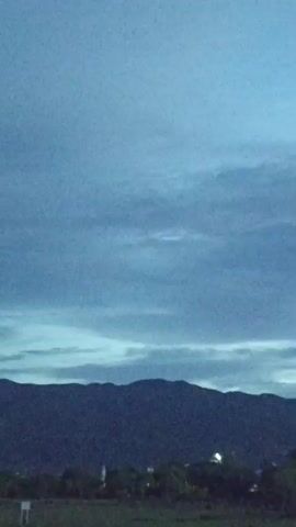 The video shows a wide shot of a natural landscape at dusk. In the foreground, there is a dark grassy field. In the middle ground, a line of trees and some distant lights are visible. In the background, a dark mountain range stretches across the horizon. The sky is a deep blue with streaks of lighter clouds, indicating either twilight or an overcast sky. The overall mood is calm and serene.