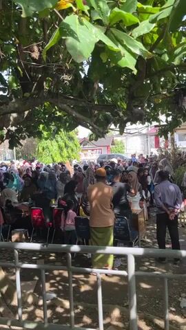 A large crowd of people are gathered outdoors, seemingly waiting for something. The foreground shows a metal fence, partially obscuring the view. Behind the fence, a large tree with broad leaves provides shade over a significant portion of the scene. Many people are dressed in casual clothing, with a notable number of women wearing headscarves. Some individuals are seated on chairs, while others stand. In the background, buildings and vehicles are visible, suggesting an urban or semi-urban environment. The overall atmosphere appears to be one of anticipation or collective engagement, with individuals interacting, conversing, and observing.