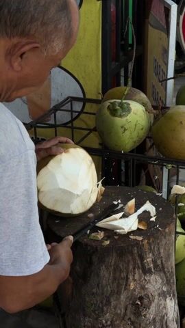 An older man, seen from a high angle behind his right shoulder, is meticulously peeling a green coconut. He is wearing a light grey t-shirt and has short dark hair. He holds the coconut firmly with his left hand on a sturdy, dark brown tree stump that serves as a cutting board. With his right hand, he skillfully uses a large knife with a dark handle and a silver blade to shave off pieces of the coconut's outer husk, revealing the lighter, smoother inner layer. Several pieces of the removed husk are scattered on the tree stump around the working area.

In the background, behind the man, more coconuts are visible, stored in black metal wire racks. These coconuts vary in color from bright green to a more yellowish-green. A yellow banner or sign is prominent, displaying the text "SATE AYAM" (chicken satay) and an image of a tall glass filled with a light brown, iced beverage. Another sign, partially visible, appears to have repetitive text, possibly related to drinks or food items. The setting suggests an outdoor street food stall or market, with general bright daylight illumination. The man appears focused and adept at his task.