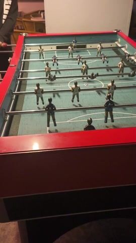 A high-angle, static shot shows a foosball table with players competing. The table has a red frame and blue-green playing surface with white markings, simulating a soccer field. The players are miniature figures, some wearing blue and yellow uniforms, others wearing white and yellow. Two people are playing, one person in a black hoodie and the other in a gray suit. The person in the hoodie is holding onto the handles on one side of the table. The players spin the rods to move the figures and hit the black ball back and forth. The environment is indoors, and a chair and other furnishings can be partially seen in the background. Upbeat music plays throughout the video.