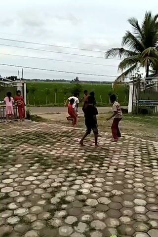Several children are playing with a soccer ball in a grassy area in front of a house. Two girls and a woman are standing near the gate to the property, watching the game. The area is paved with cobblestones in a honeycomb pattern. A green rice field is visible in the background, separated from the houses by a paved road. The sky is overcast. A tall palm tree is to the right of the group of children playing soccer.