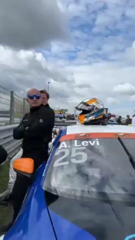 The camera pans across a group of four men standing near a race car at a race track. The race car is blue and white with the number 25 and the name "A. Levi" displayed on the windshield. An orange and blue racing helmet sits on the roof of the car. The man closest to the camera is bald and wearing sunglasses and a black jacket. He has his arms crossed. Next to him is a man in a white and blue racing suit. Another man in a black jacket and pants stands behind them. The fourth man is partially visible to the left. A metal guardrail runs along the edge of the track, and there are other race cars and people in the background. The sky is cloudy with patches of blue.