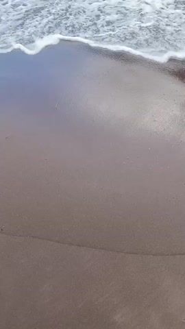 A slow-motion shot of gentle waves lapping onto a dark sandy beach. The water is foamy and white as it recedes, leaving behind a wet, reflective surface on the sand.