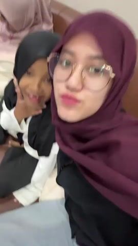 The video features two women and a young girl sitting on a brown sofa indoors. A woman in a maroon hijab, glasses, and braces is holding the camera to record a selfie video. She smiles at the camera throughout the clip. Beside her is a young girl wearing a black hijab and a white top. The girl initially poses for the camera with a slight smile, then turns her head away, covers her mouth, and laughs heartily, throwing her head back. Another woman, wearing a light pink/cream hijab, is partially visible in the background, also seated on the sofa. The scene depicts a joyful and relaxed interaction, likely in a living room, with a light-colored, textured wall behind them.