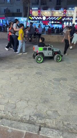 At night, a young child drives a gray toy Jeep with green accents on a cobblestone path in a public area. The child, wearing a black shirt, sits in the vehicle and operates it. Pedestrians walk past the Jeep, and several buildings, visible in the background, are illuminated with various lights and signs. A man in a maroon shirt and gray pants stands nearby, watching the child. The path appears to be a pedestrian zone, and the scene is bustling with people enjoying the evening.