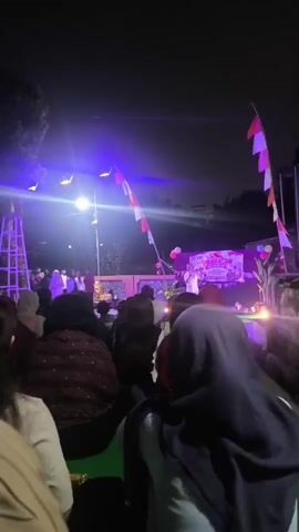 The video begins with a wide shot of an outdoor event at night. There is a stage illuminated by purple and white lights, with a banner that is unreadable due to the blurriness. Several people are gathered in front of the stage, facing it. Indonesian flags are visible on poles. The camera then cuts to a series of blurry shots of people's faces and torsos. One shot focuses on a woman with glasses making a funny face, while another shows a woman sticking her tongue out. The remainder of the video consists of very blurry, fast-moving shots of people. The overall impression is of a casual gathering, possibly a celebration or performance, with attendees taking playful photos and videos.
