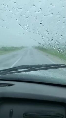 The video shows a view from inside a car driving on a wet road in the rain. Raindrops are visible on the windshield, distorting the view of the road ahead. The windshield wipers are in motion, intermittently clearing the view. The road appears to be a two-lane highway with green vegetation on either side. The sky is overcast and the overall lighting is dim, indicative of a rainy day. The interior of the car is visible at the bottom of the frame, including a dashboard and a portion of a seat. The video appears to be a first-person perspective of someone driving under rainy conditions.