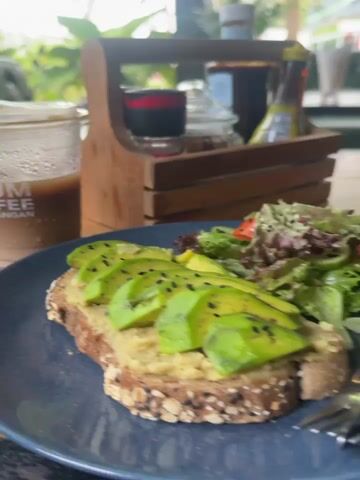 The video shows a table setting with a partially eaten avocado toast, a side salad, a plastic cup of iced coffee, and a wooden caddy containing various condiments. The avocado toast is on a dark blue plate and features slices of avocado sprinkled with black sesame seeds. The salad includes lettuce and tomato. The coffee cup has the logo "NUUM COFFEE KOH PHANGAN." The wooden caddy contains a spice grinder, a small jar, and bottles of soy sauce and what appears to be sriracha. The table is wooden and appears to be outdoors or in an open-air restaurant, as greenery is visible in the background. A leather couch and a wooden chair are also partially visible.