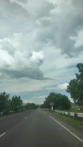 The video shows a forward-facing view from a vehicle traveling on a straight, paved road in Horom, Armenia. The road has two lanes, separated by a white line. On either side of the road, there are trees and vegetation. In the distance, the road stretches towards a horizon line marked by a soft, hazy mountain range under a vast sky.

The sky is dramatic, filled with large, dark, and white cumulonimbus clouds, suggesting imminent or recent rainfall, or simply a stormy weather pattern. Despite the clouds, there are patches of blue sky visible, and the overall lighting suggests it is daytime, possibly late morning or afternoon, given the brightness.

A white guardrail runs along the right side of the road, bordering a grassy area. On the left side, there are trees and some scattered structures, including what appear to be utility poles and possibly a distant antenna tower. A blue sign with white lettering and a circular logo is visible on the right side of the road, mounted on a post. While the text on the sign is not entirely legible due to the distance and resolution, it appears to be informational, possibly a road sign or a local announcement.

The scene conveys a sense of journey and the vastness of the landscape. The weather contributes to a somewhat dramatic and moody atmosphere, contrasting with the otherwise calm and linear progression of the road. There are no people or other vehicles directly visible in the frame, emphasizing the solitary nature of the drive.