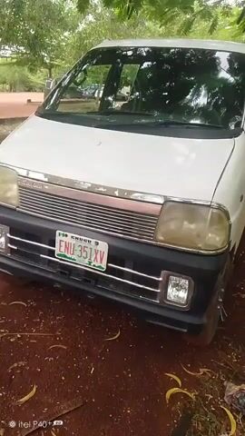 This media file displays a white minivan parked outdoors, with a focus on its front end. The vehicle has a license plate that reads "ENU 351XV" and appears to be a Nigerian plate, as indicated by "ENU" which likely refers to Enugu State. The surrounding environment suggests a rural or semi-rural setting, with trees and foliage visible, casting shadows that hint at daylight conditions. The ground is covered in dirt and fallen leaves, with some grass on the side. In the background, other vehicles and a few people can be seen, indicating some level of activity or public space. The overall impression is of a still-life shot of the vehicle in its natural environment, possibly in a parking area or along a road.
