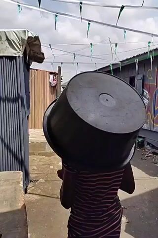 The video shows a person carrying a large metal pot on their head while walking down a narrow alleyway. The alleyway is lined with wooden buildings, some of which have graffiti on them. There are green and white pennants strung across the alleyway. Several people are walking in the same direction as the person carrying the pot.  The ground is uneven and dusty. Clothes are hung outside a building on the left of the frame.