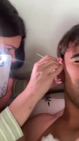In an indoor setting, possibly a bedroom, a woman with dark hair is assisting a man with an ear cleaning. The woman, partially visible on the left, holds a white cell phone with its flashlight on, illuminating the man's ear. She is wearing a light green striped top and has a small tattoo on her wrist. With her right hand, she uses a metallic ear pick to remove earwax from the man's right ear. The man, with brown hair and light stubble, is shirtless and lies on a pillow with a leaf pattern. He initially winks and makes a slightly uncomfortable expression, then smiles at the camera. A piece of cotton wool, stained with yellowish-brown earwax, rests on his chest, indicating the results of the cleaning. The woman then takes the ear pick and displays the collected earwax on the cotton. The overall mood appears casual and slightly humorous, with the man reacting playfully to the process. No visible text is clearly legible on the phone.