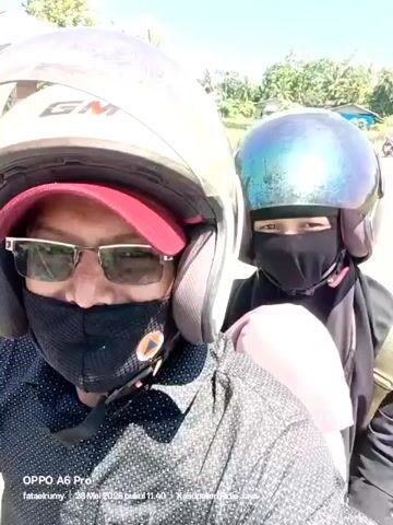 Two people are riding on a motorcycle. The person in front is wearing a helmet, sunglasses, a red baseball cap, and a black face mask. The person behind is also wearing a helmet and a black face mask. The background shows a road and some trees under a bright blue sky. The phone's timestamp indicates the video was recorded on May 28, 2026, at 11:40 AM.