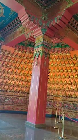 The video shows the interior of a Buddhist temple. The walls are covered in numerous small golden Buddha statues arranged in a grid pattern against an orange background. In the center of the frame, a large golden Buddha statue is seated on a decorative altar, adorned with flowers. The ceiling is intricately painted with a Dharma wheel symbol. A large red pillar stands in the foreground, with ornate carvings and decorations on the ceiling and walls. Velvet ropes and stanchions are visible, suggesting a designated viewing area for visitors. The floor appears to be polished marble.