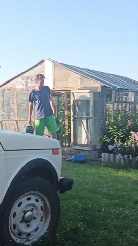 A young person wearing a blue t-shirt and green shorts waters a plant with a blue watering can in front of a greenhouse. A white car is parked in the foreground. The scene is outdoors in a grassy area under a clear blue sky.