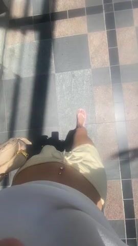 The video, taken from a high angle, shows a person walking on a paved surface. The surface is composed of square and rectangular tiles in varying shades of grey, beige, and black. The person's bare stomach is visible, adorned with a navel piercing. They are wearing a white top, light green shorts with a drawstring, and pink flip-flops. A small, beige crossbody bag is visible on their left hip. Long shadows stretch across the ground, suggesting bright sunlight. The person is moving forward, their feet visible as they step onto the tiled surface.