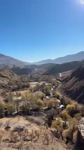 The video shows a wide valley with a river running through it, viewed from a high vantage point. Mountains are visible in the background under a clear blue sky with the sun shining brightly. The valley is sparsely populated with trees, many showing autumn colors. A road winds through the valley. A person wearing a striped shirt and jeans walks into the frame and stands at the edge of the cliff, looking out over the valley. They raise a camera or phone to take a picture.