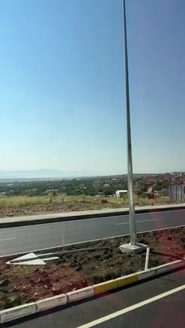 The video shows a view from a moving vehicle looking out over a semi-urban landscape under a clear blue sky. In the foreground, there is a paved road with white lane markings and a concrete curb with yellow and white stripes. To the left of the road is a dirt and gravel embankment with sparse vegetation. Further into the scene, there is a residential area with scattered houses, trees, and green spaces. In the distance, a hazy mountain range is visible on the horizon. There are several poles, including what appear to be lampposts, and road signs are also present. The camera appears to be looking out a window, as indicated by the reflection visible in the bottom portion of the frame. The overall impression is of a sunny day and a journey through a suburban or rural area.