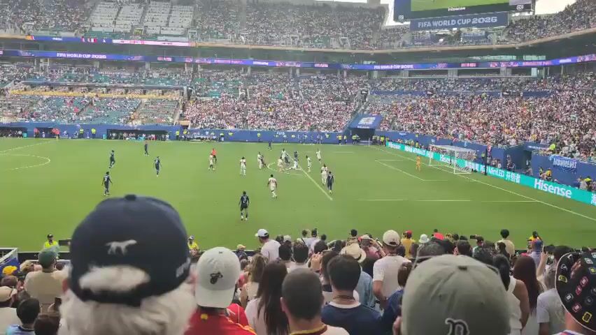The video shows a wide shot of a soccer game taking place in a crowded stadium. The field is green with white lines. There are many players on the field, some in blue uniforms and others in white. The stands are filled with spectators. The video is shot from behind a section of the crowd, with the backs of people's heads and shoulders visible in the foreground. Some people are wearing hats and casual clothing. The stadium has multiple tiers of seating and banners are visible in the background. The overall scene depicts a lively sporting event.