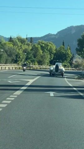 A motorcycle is seen on the left side of the road, moving away from the camera. On the right side of the road, a white vintage car is turning a corner. The car has a spare tire mounted on the back. The road is paved and marked with white lines. Trees and hills are visible in the background under a clear blue sky.