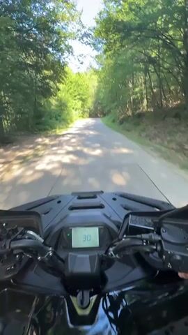 This first-person perspective video captures a thrilling ATV ride through a lush, tree-lined road in Orlovtsi, Bulgaria. The bright sunlight dappling through the leaves indicates a clear, warm day, likely late morning or early afternoon. The ATV's digital dashboard is visible, displaying a speed of 32, then 35, and finally 37, suggesting the rider is accelerating. The road is paved but appears somewhat narrow, with dense green foliage flanking both sides, creating a tunnel-like effect. The trees are tall and leafy, characteristic of a forest or wooded area.

The focus of the video is the experience of riding the ATV, with the handlebars and dashboard prominently featured in the foreground. A hand is visible on the right handlebar, operating the controls. The dynamic blur of the trees and the road conveys a sense of speed and movement. The overall mood is one of exhilaration and enjoyment of the natural surroundings. No people other than the rider are visible, but the clear sky and vibrant greenery suggest a pleasant outdoor adventure. The visual perspective is immersive, placing the viewer directly into the driver's seat.
