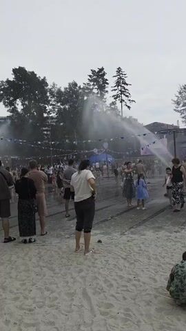 The video shows a group of people gathered in an outdoor area, possibly a park or a festival, on a cloudy day. A fine mist of water is being sprayed into the air from multiple sprinklers, creating a hazy atmosphere. Some people are standing and observing, while others appear to be walking through the mist. A few children are visible, and some adults are interacting with them. There are trees and some structures in the background, including what looks like a stage or a booth with lighting. A large orange vehicle, possibly a truck or a utility vehicle, is partially visible on the right side of the frame. The ground appears to be a mixture of sand and paved surfaces. The overall impression is that of a casual outdoor event where water is being used for cooling or as part of an attraction.