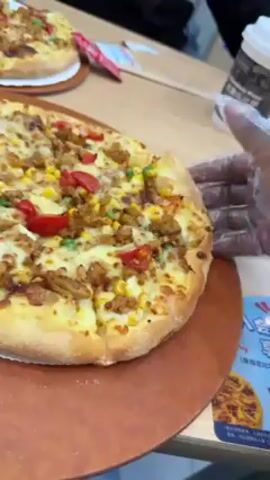 Here's a description of the video:

A gloved hand picks up a slice of pizza from a pizza sitting on a wooden-style serving board. The pizza has a light-colored crust and is topped with cheese, what appears to be shredded chicken, corn kernels, diced red peppers, and green peas. Two other pizzas are partially visible in the background, and a cup with a lid is on the table. The person appears to be wearing a black sweatshirt. A sign with pizza graphics and text is visible on the right edge of the frame. The environment looks like a restaurant or fast food establishment.
