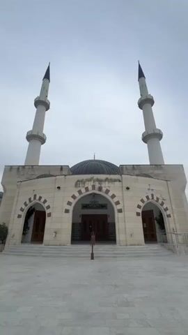 The video shows a mosque with two minarets and a dome. The exterior is made of light-colored stone. There are arched doorways and windows. In the foreground, there is a paved courtyard with a fountain in the center. Several people are seated at tables near the fountain. Mountains are visible in the background. The sky is overcast.