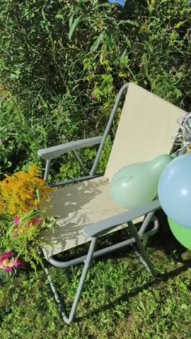 The video shows a picnic scene in a grassy outdoor area. It starts with a shot of a folding chair adorned with a bouquet of flowers. Then, a black bag is seen on the grass. The camera then moves to focus on a metal barbecue grill with glowing coals and flames, placed on a mat on the lawn. The surrounding environment is lush green with trees and foliage in the background, suggesting a sunny day.