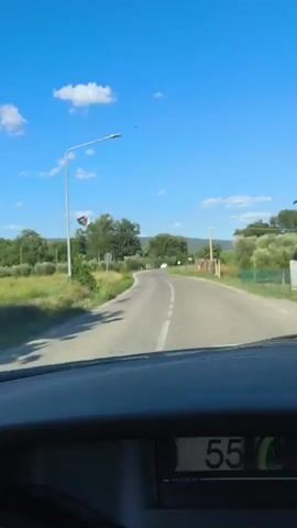 The video shows a view from inside a vehicle traveling on a paved road through a rural area. The road curves to the right and appears to be surrounded by vegetation, including trees and fields of what look like olive groves. The sky is blue with scattered white clouds. A streetlamp with a piece of paper attached to it is visible on the left side of the road. In the distance, there are a few trees and possibly some buildings. The dashboard of the vehicle is visible in the foreground, with a digital display showing "55". There are no people clearly visible in the video. The overall impression is one of a scenic drive on a sunny day.