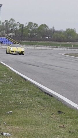 The video shows a yellow convertible Porsche driving on an asphalt track. The car has a black stripe running down the middle of the hood, and the license plate is visible. There are grass fields on either side of the track and signs are visible in the background, one which says "RACING BAZAR" and another that says "MA-RI". The weather appears overcast and the road is dry.