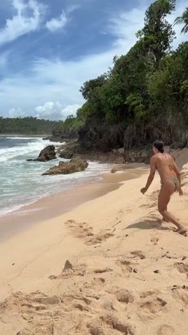 The video shows a woman on a beach. She jumps in the air and then kneels to pick up a handful of sand. She throws the sand over her shoulder, turns, and walks toward the water. The beach has pale sand and is bordered by rocks and green vegetation on one side and the ocean on the other. The sky is blue with scattered clouds.