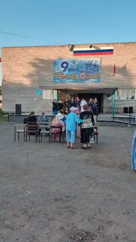 At the 00:00 mark, a group of individuals are gathered outside a building. The building has a large banner above its entrance that reads "9 MAY" and features imagery related to Victory Day. Several people are seated on chairs facing a group of performers who are standing on a raised platform in front of the building. Two women in the foreground are standing and appear to be interacting. The environment appears to be outdoors, with a clear sky and what looks like a gravel or dirt ground. The lighting suggests it is daytime, likely in the late afternoon or early evening.