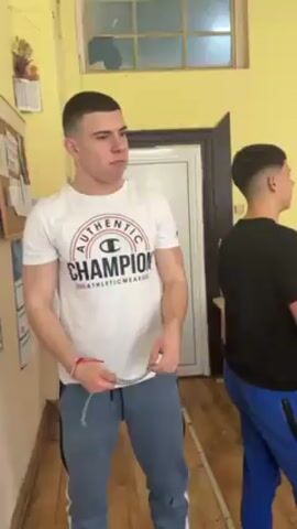 A young man in a white Champion t-shirt and blue sweatpants stands in a school hallway, examining a chain in his hand. He looks up at a bulletin board on the yellow wall to his left. Another young man wearing a black t-shirt and blue sweatpants stands to the right, partially obscured. The hallway features a window and a door visible in the background.