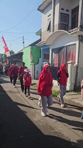 A group of people are walking down a street, decorated with red and white flags. The people are mostly wearing red shirts and some are wearing red headscarves. The street is narrow and lined with houses and some vegetation. The sky is clear and blue. The sun is shining, casting shadows on the street. The people appear to be participating in some kind of procession or celebration. There is a cart in the background with people on it.