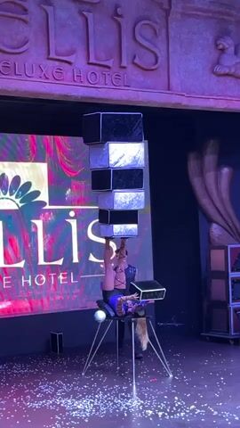A performer is balancing a tower of boxes on her feet while in an inverted position. She is on a stage in front of a backdrop with the words "ELLIS DELUXE HOTEL" visible. There are confetti scattered on the stage floor, and another person is standing to the left of the performer. The performer is wearing a blue and black outfit. The boxes are stacked vertically, alternating in color between black and silver. The performer's legs are bent, and her feet are supporting the bottom box of the tower.