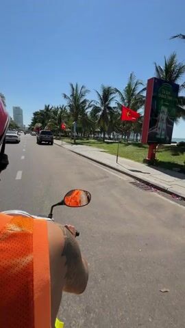 A first-person perspective video shows a person riding a scooter on a paved road. The scooter has an orange handlebar and a reflective mirror. The rider is wearing an orange vest and has a tattoo on their arm. On the right side of the road, there is a green lawn with numerous palm trees. In the background, there is a body of water, possibly the ocean, and a clear blue sky. A dark SUV is visible parked on the left side of the road, with a red flag on a pole nearby. Several motorcycles are parked on the grass. The overall scene depicts a sunny day at a tropical or coastal location.