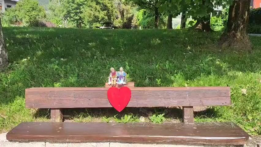 The video shows a weathered wooden bench in a park. Two small figurines of an elderly couple are sitting on the bench, facing forward. In front of them, attached to the bench's backrest, is a large red heart decoration. The bench is positioned on a grassy area with trees in the background. The lighting suggests it is daytime, with sunlight dappling the scene.