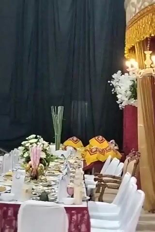 A long table is set for a formal event. The table is covered with a white tablecloth and decorated with a maroon runner. White chair covers are placed on the chairs around the table. The table is laden with plates of food, silverware, glasses, and decorative items. In the center of the table, there is a floral arrangement with white flowers and some pink decorative elements, as well as a basket of fruit. Tall, thin green stalks are arranged behind the floral centerpiece. The background consists of dark fabric drapes. The lighting is dim, suggesting an indoor event.