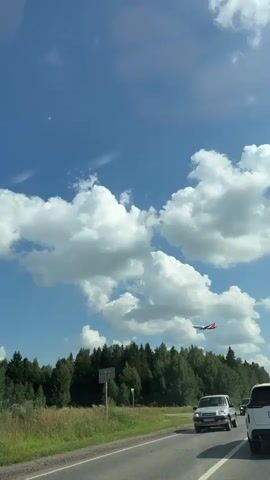The video captures a view from a moving vehicle, likely a car, on a sunny day with a blue sky and scattered white clouds. A silver SUV is visible in the foreground on the left side of the road. Further down the road, another dark-colored vehicle is seen. In the distance, a line of lush green trees forms the horizon. A red and white airplane is flying across the sky above the trees. The overall scene suggests a rural or suburban setting during daylight hours.
