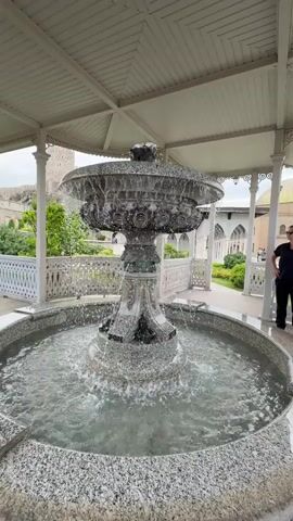 A ornate, multi-tiered fountain with water flowing from the top tier cascades into a circular basin. The fountain is made of a light-colored stone, possibly marble or granite, with intricate carvings and metallic accents. The fountain is situated under a white, slatted roof supported by columns. To the right of the fountain, a person wearing a black t-shirt and black pants stands looking towards the fountain. In the background, a white ornate railing runs along a walkway. Beyond the railing, there is greenery and a building with a domed roof. To the far left, a stone wall is visible. The overall environment appears to be an outdoor courtyard or garden area.