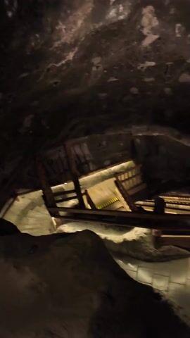 The video shows a group of people on a guided tour in a dimly lit cave. They are standing on a wooden platform with stairs leading down. A tour guide is pointing towards a large rock formation in the cave. The cave walls are dark and uneven.